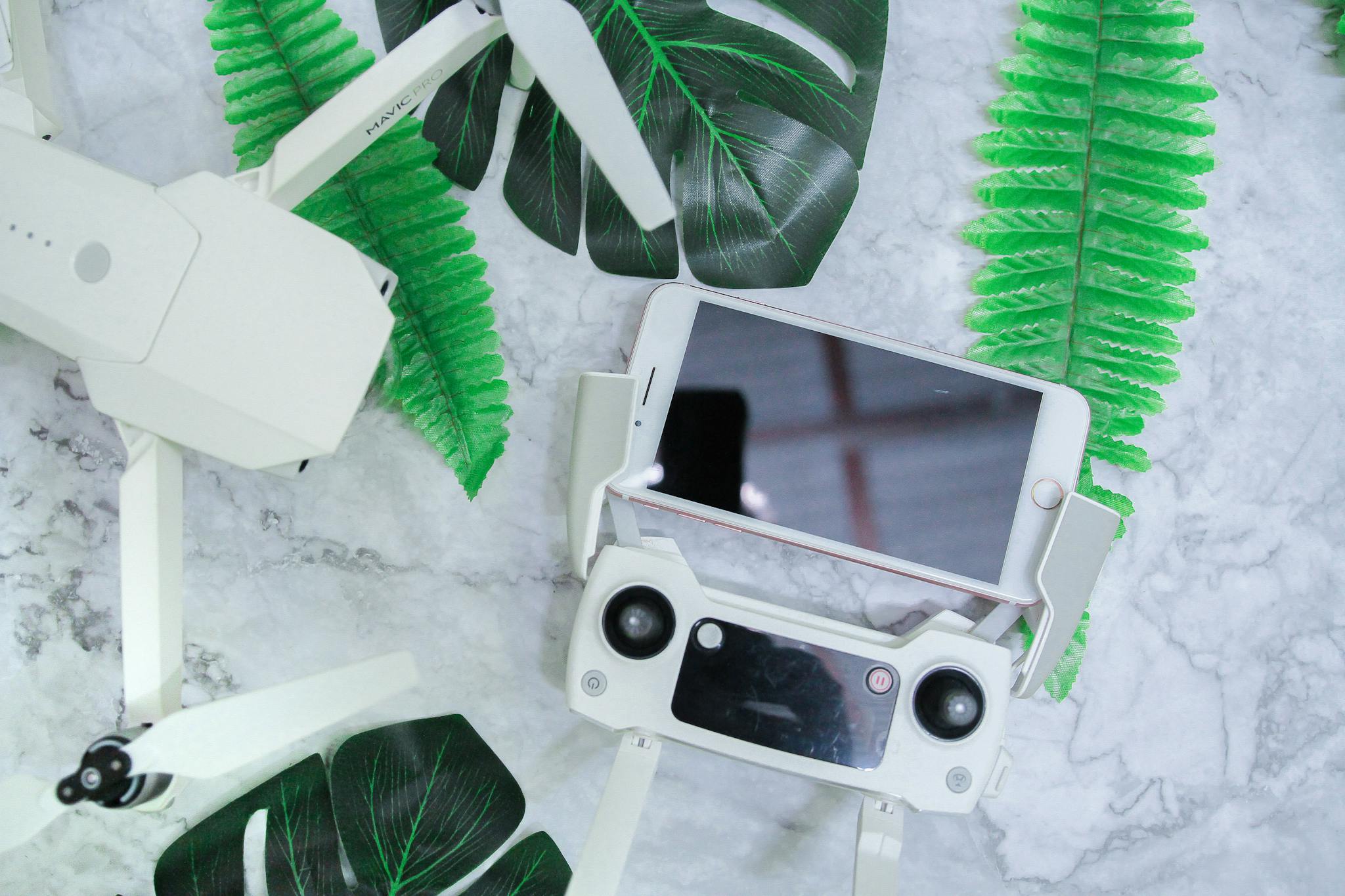 From above of black screen of smartphone placed above flight controller near drone on desk decorated with tropical plants during daylight